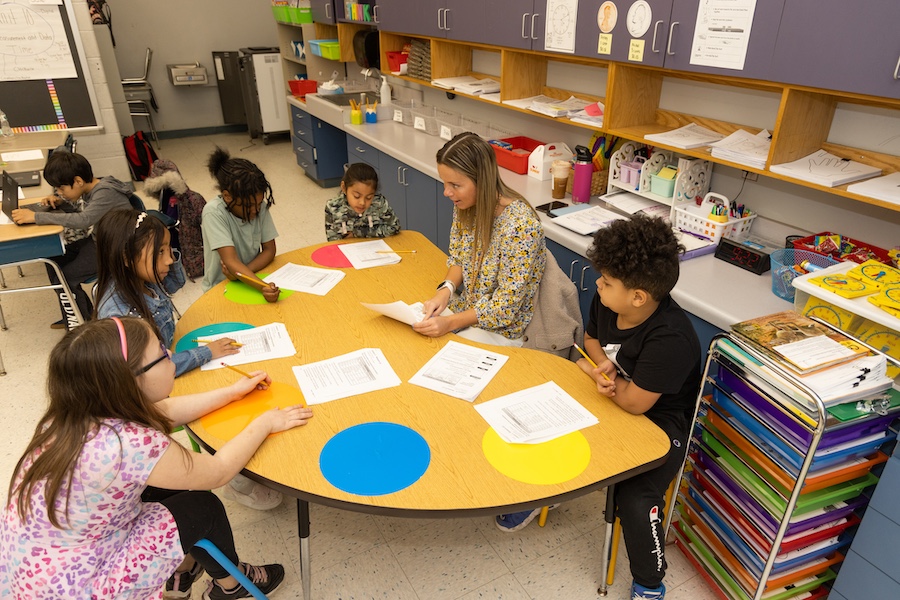 A student teacher sitting at a semi-circle table with 5 elementary level students, working on a worksheet with them. 